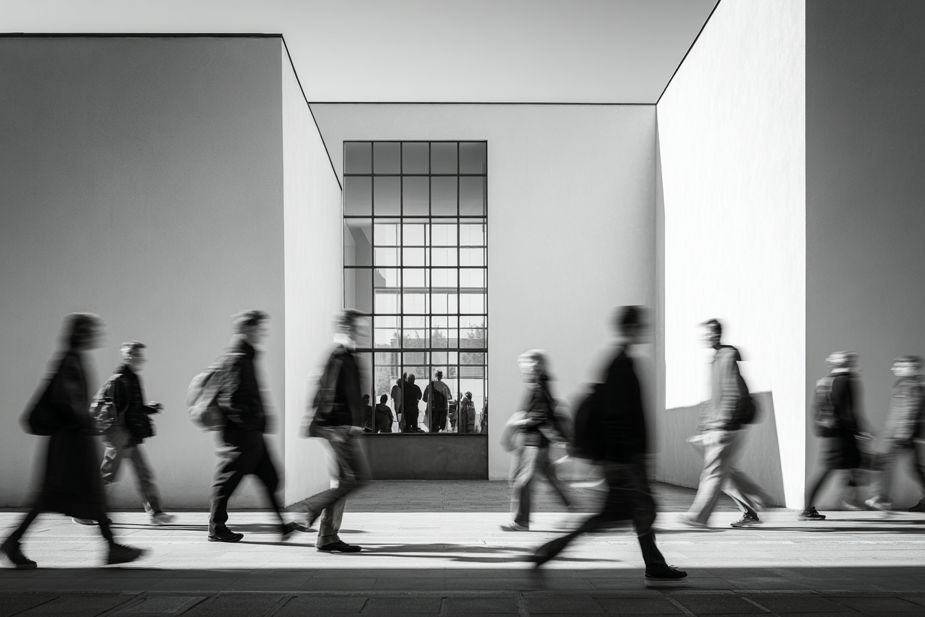 People walking past Bauhaus-style white walls and glass windows, minimalist architecture with soft directional light and shadow emphasizing motion and form.